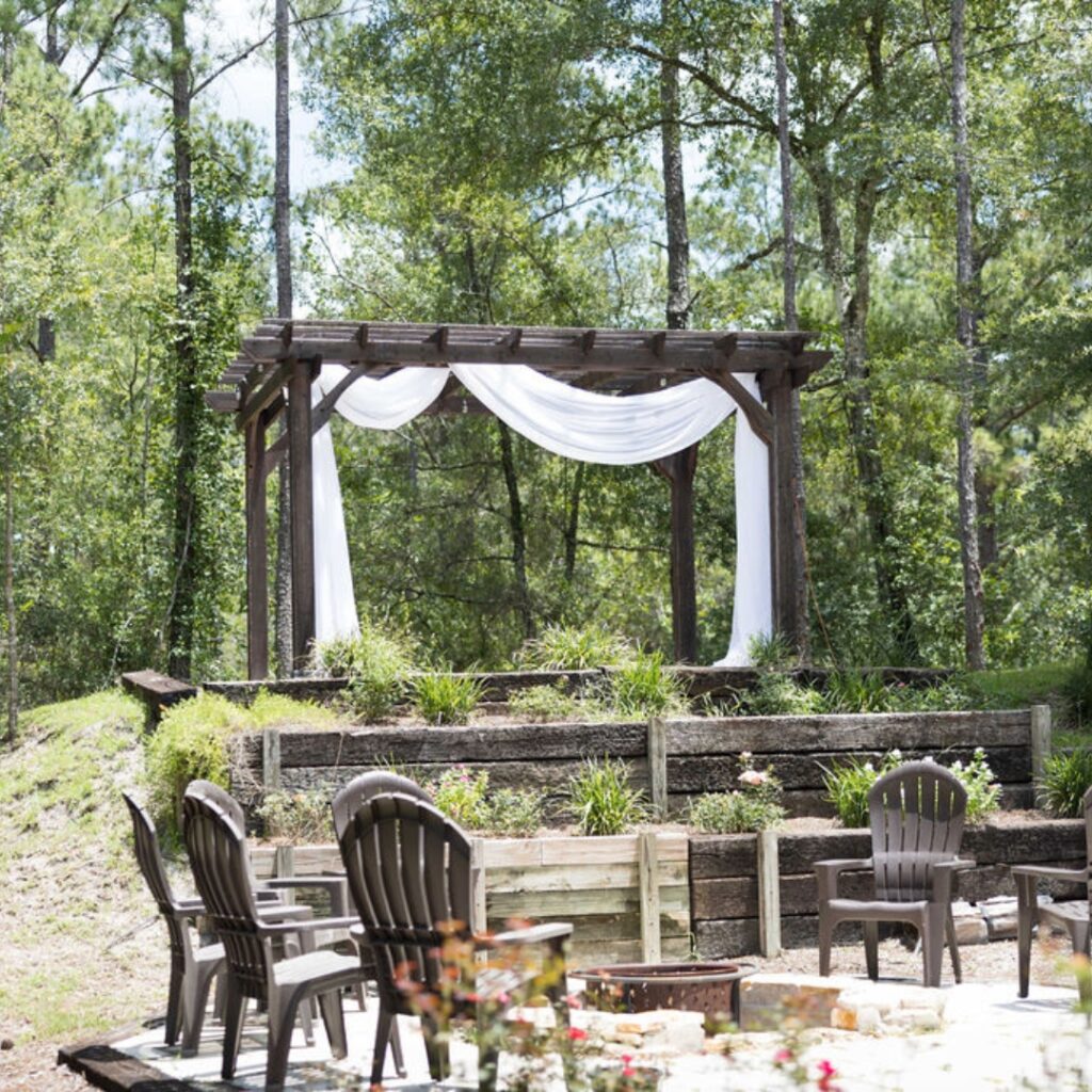 image of a small wooden pergola draped in white draping with a fire pit and chairs in the foreground