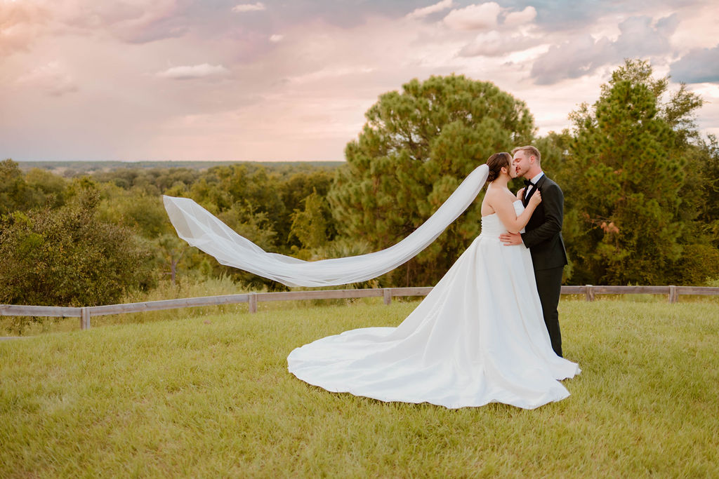 image of bride and groom hugging, bride's veil is blowing in the wind, backdrop is beautiful tree scenery